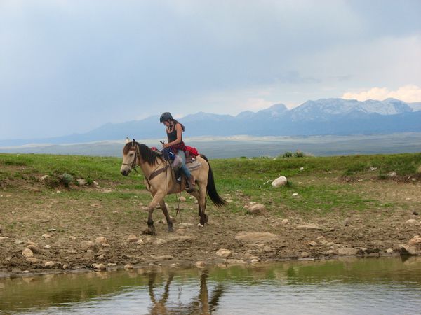 Horse packing student rides along the water's edge with mountains in the background. 