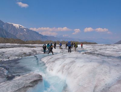 Students cross a glacier river in Alaska with towering snow-capped peaks in the distance. 