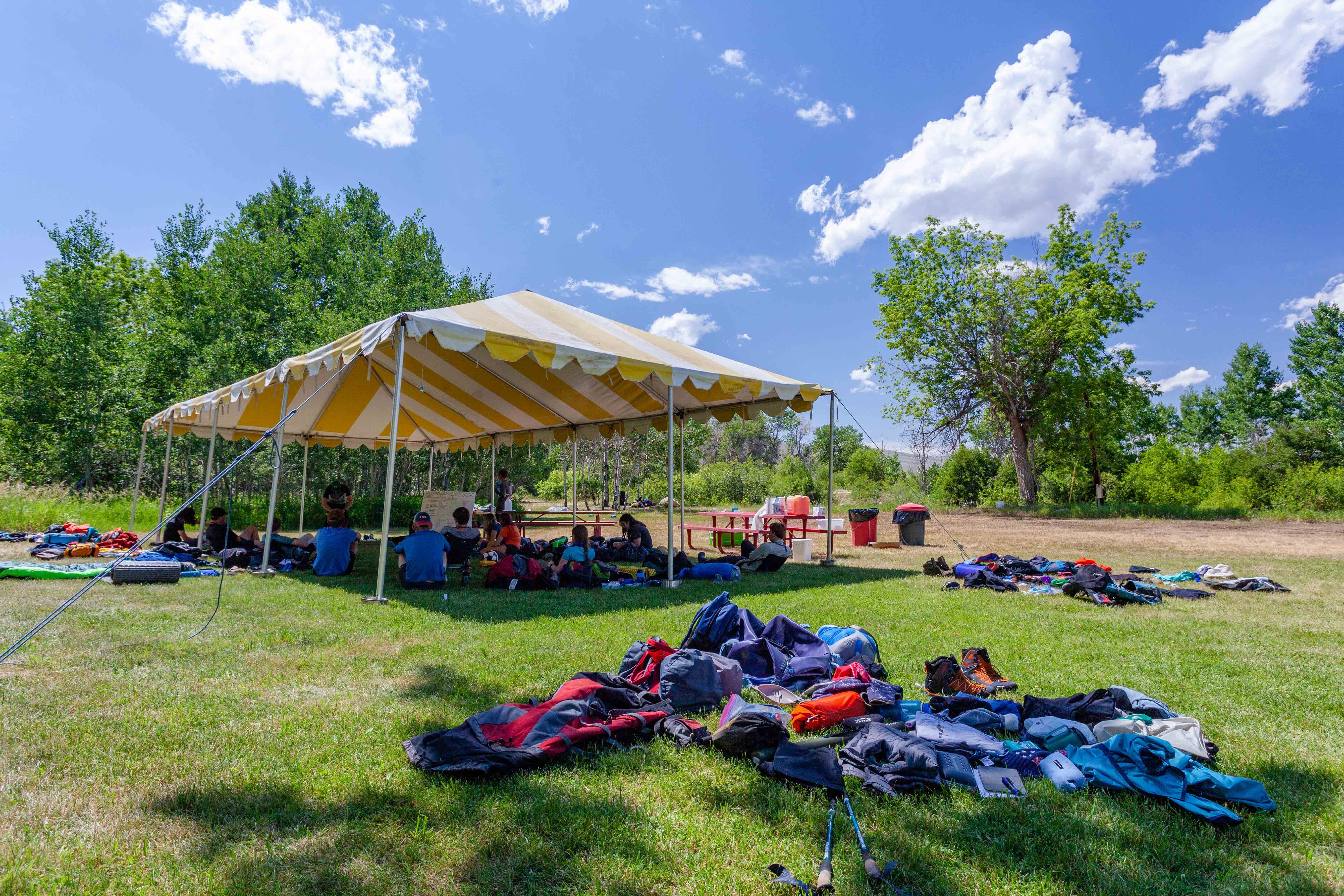Students sit under a canopy with all of their bags unpacked as they listen to an instructor.