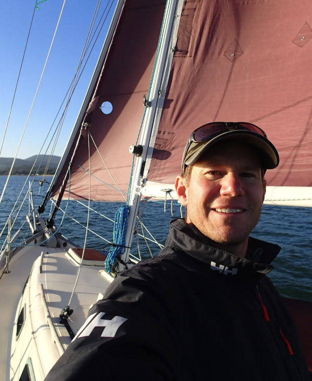 Nick Braun on a sailboat in Greece, red sail and blue seas behind him
