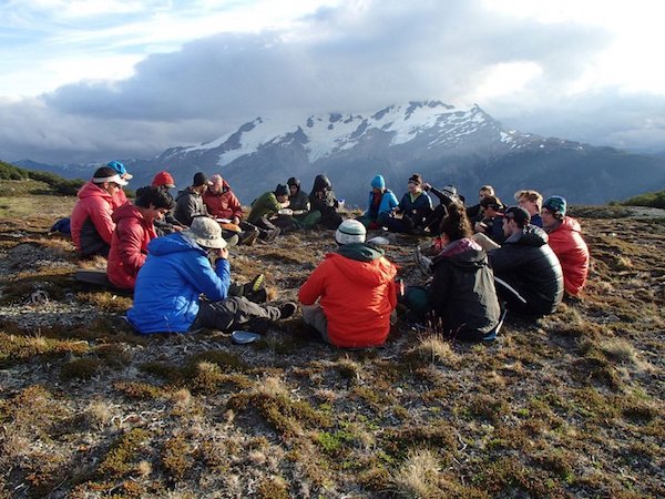 nols students gathered in a circle on the ground with snow-capped mountains in the backdrop