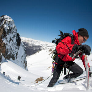 a man wearing a red coat and black snowpants, mountaineering up the side of a steep snow covered incline