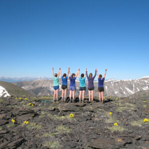 6 girls wearing various blue tops and dark shorts viewed from the back with their hands in the air looking off towards mountains
