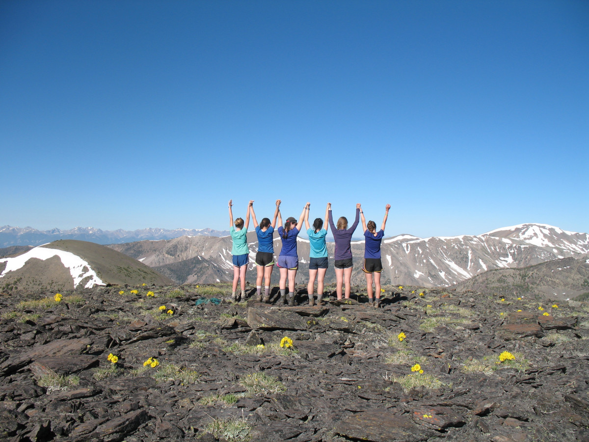6 girls wearing various blue tops and dark shorts viewed from the back with their hands in the air looking off towards mountains