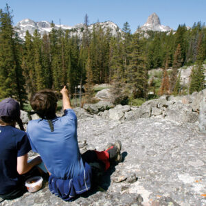 two teens facing away sitting on a rock, the one on the right pointing towards something far off into the trees in front of them