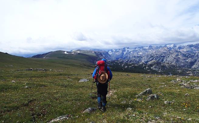 Backpacking student with trekking poles hikes across meadow toward rocky peaks in the Wind River Range.