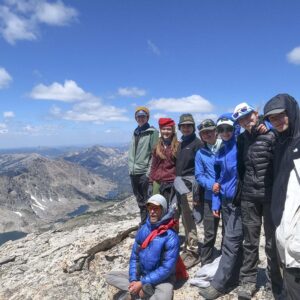 Group of teens standing on top of a large rock at elevation with mountains off in the background
