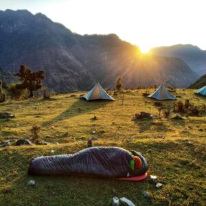 Sunrise over the mountains putting light on camp with someone in a sleeping bag on the grass out front of tents.