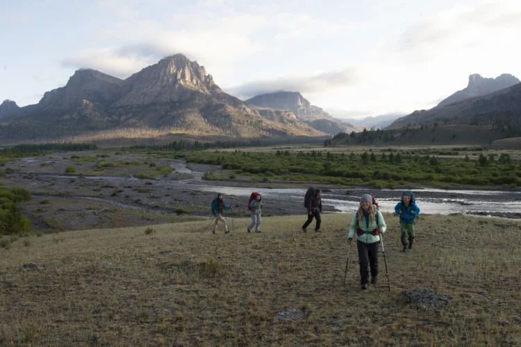 five backpackers hikeup a grassy hill. there are tall granite peaks in the background.