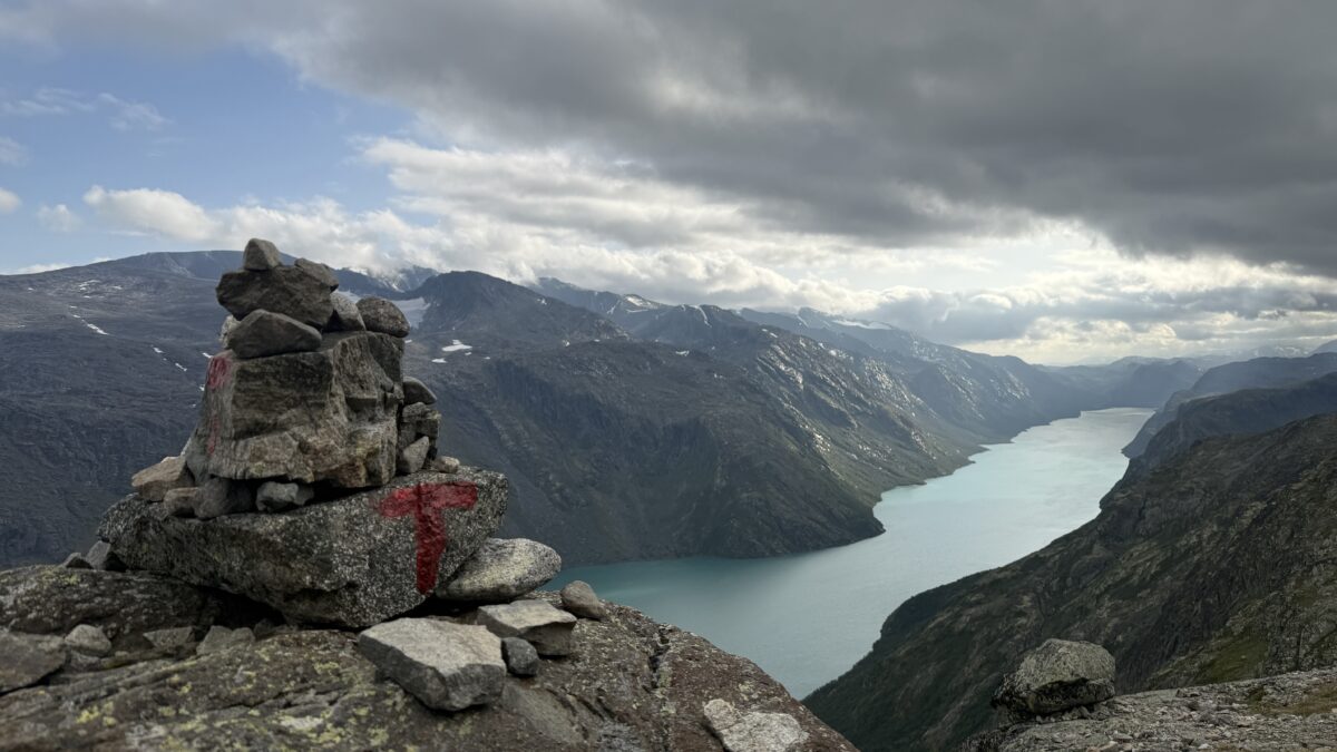 Mountains surround narrow lake with dark cloud over much of the sky and large cairn in bottom left corner.