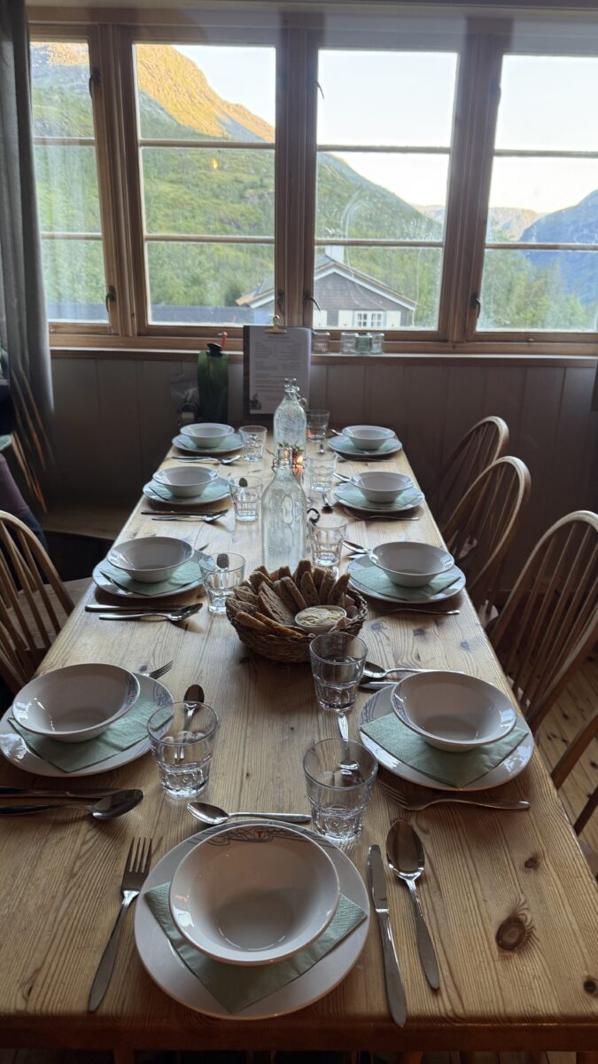 Wooden dinner table by a bank of windows set for a meal for 9 people with bowls plates, and bread.