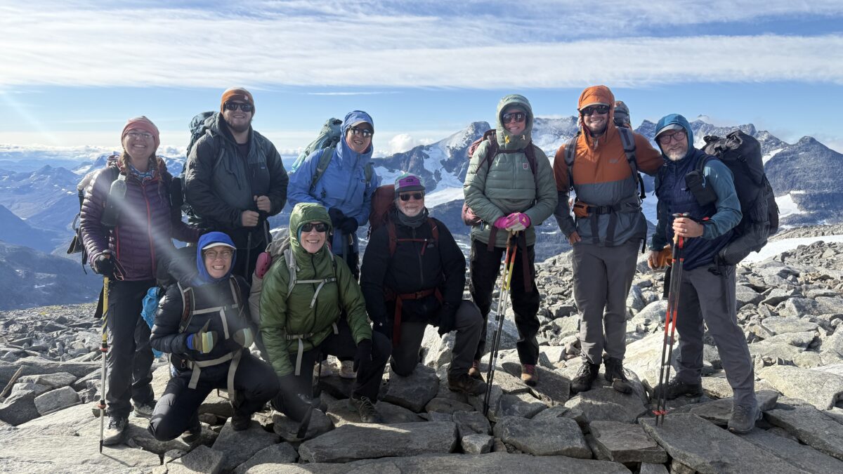 Nine members of the Norway course, dresses in puffy coats, headwear and gloves, gather on a rocky mountain side for a group photo.
