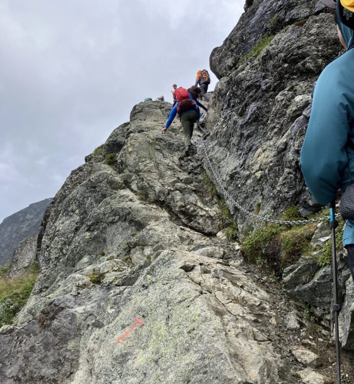 Three people hike up a steep cliff, holding onto a chain that runs along the inside of the cliff.