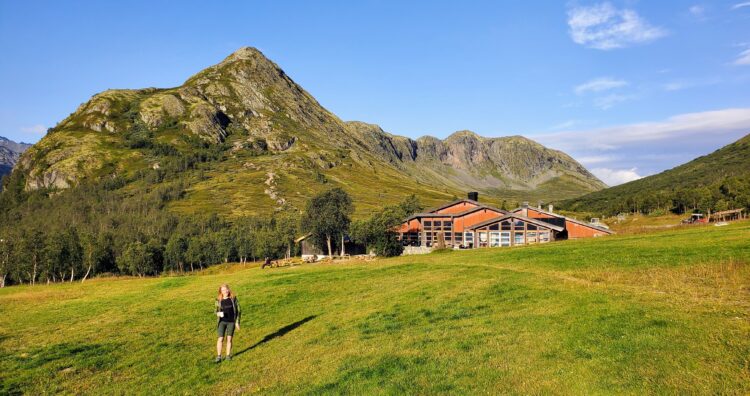 A person stands in grassy field. There is a tall rocky mountain behind her and a lodge to her right. The sky is bright blue.