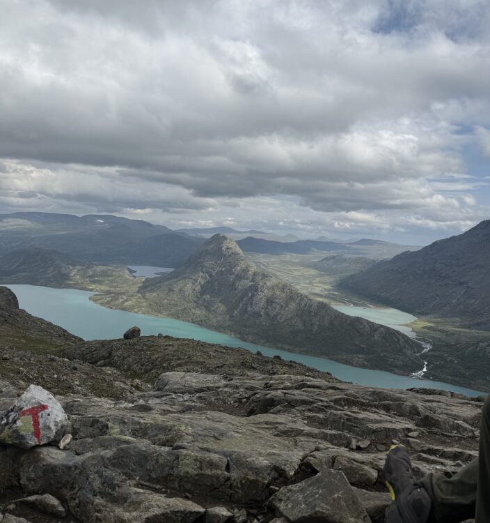 The sky is cloudy. In the foreground is a rock with a T painted on it. In the background is a turquoise lake and steep mountains.
