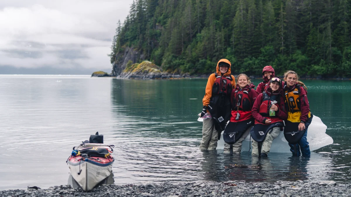 A group of four girls wearing PFDs and paddling clothes stand in ankle-deep water in front of an iceberg. There is a sea kayak on land on the left.