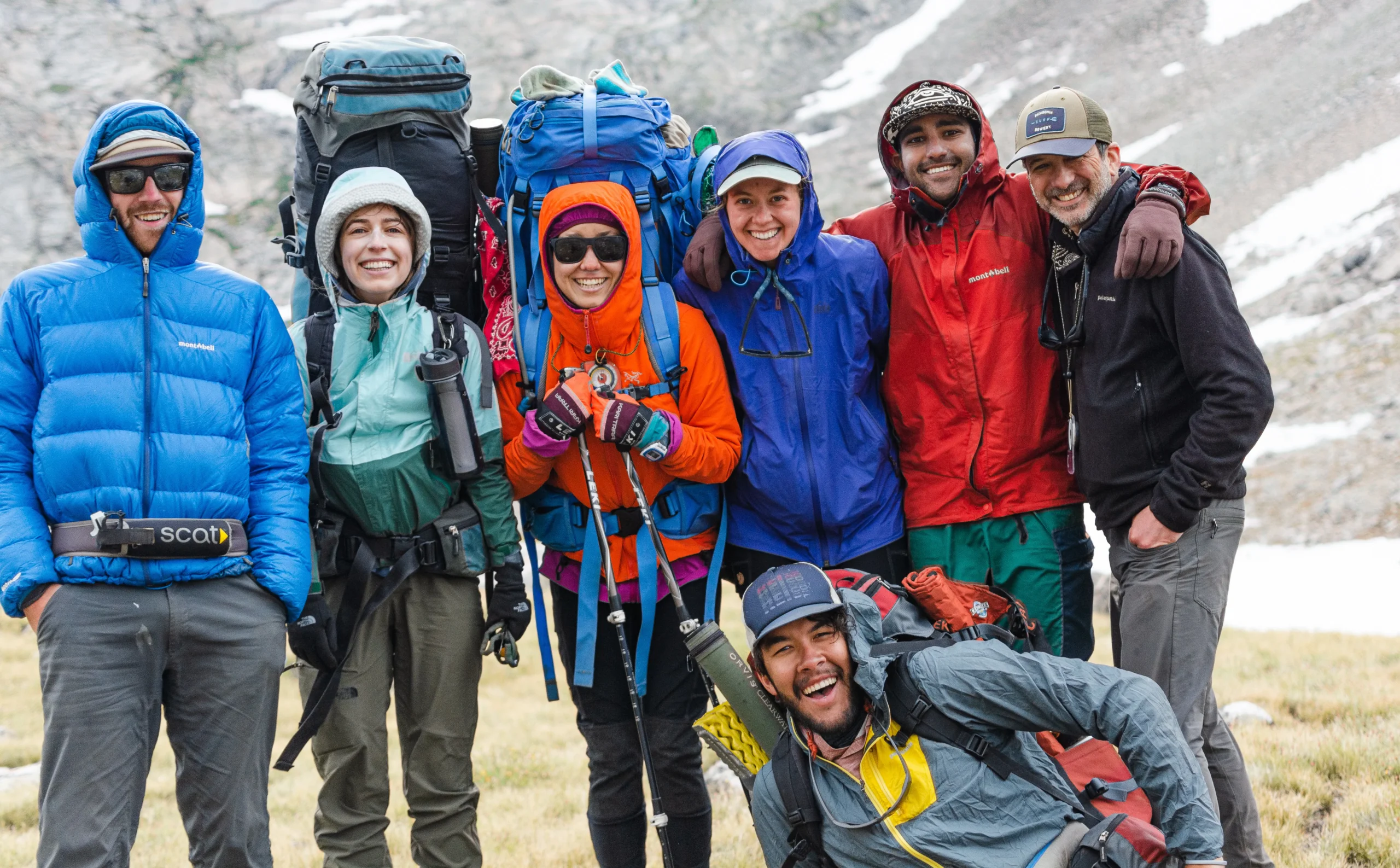 7 people wearing jackets and backpacks pose together at the base of a mountain.