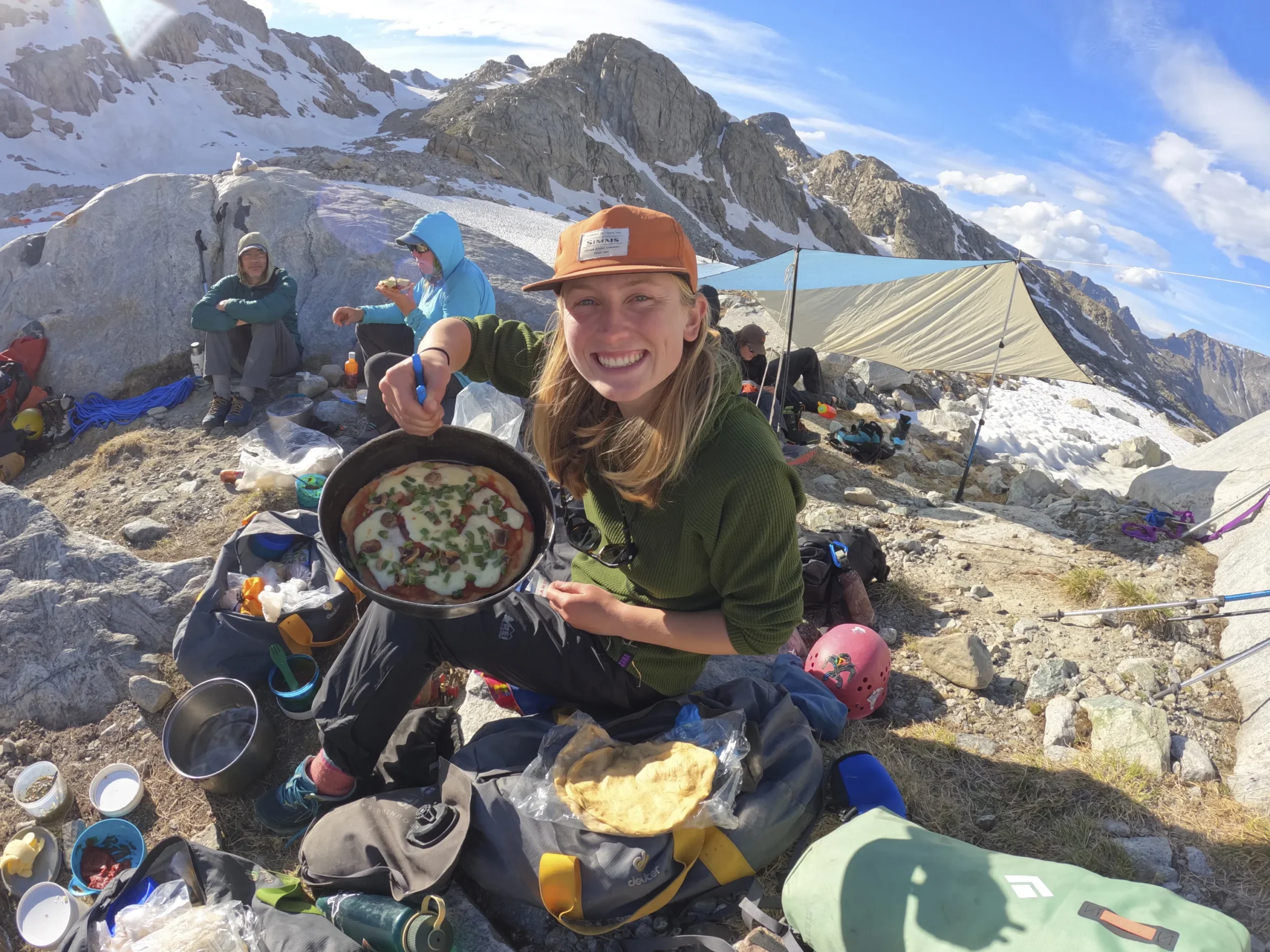 A girl holds up a pizza in a fry pan. She is sitting on the ground, surrounded by granite peaks.