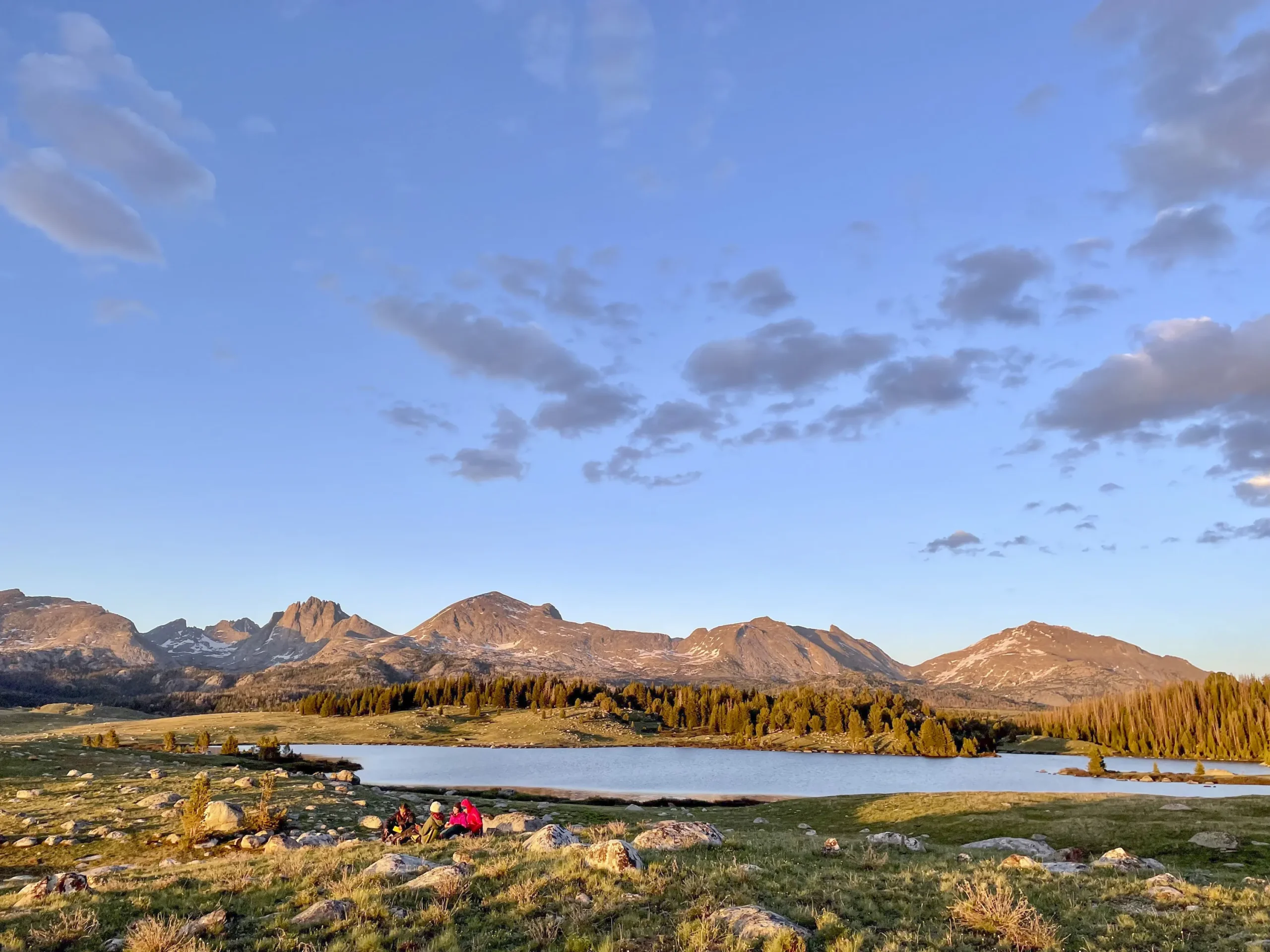 A range of granite peaks in the alpenglow. There is a lake and a small group of people in the foreground.