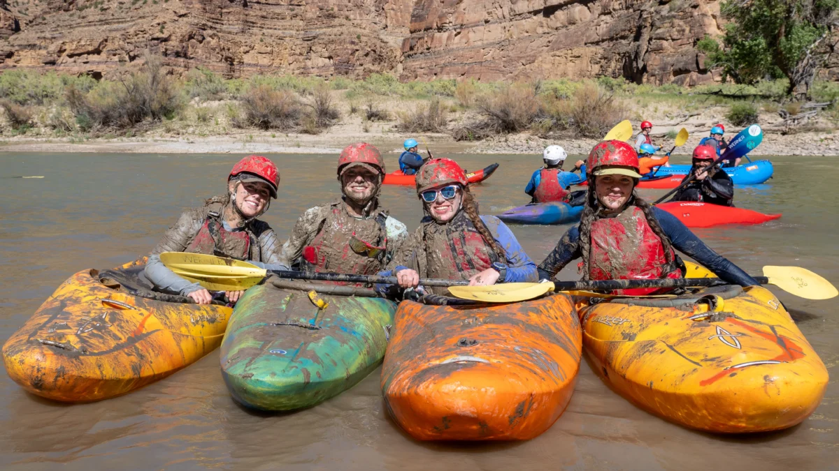 Four girls sitting in river kayaks and wearing red helmets and sunglasses pose together, smiling. They are covered in red mud