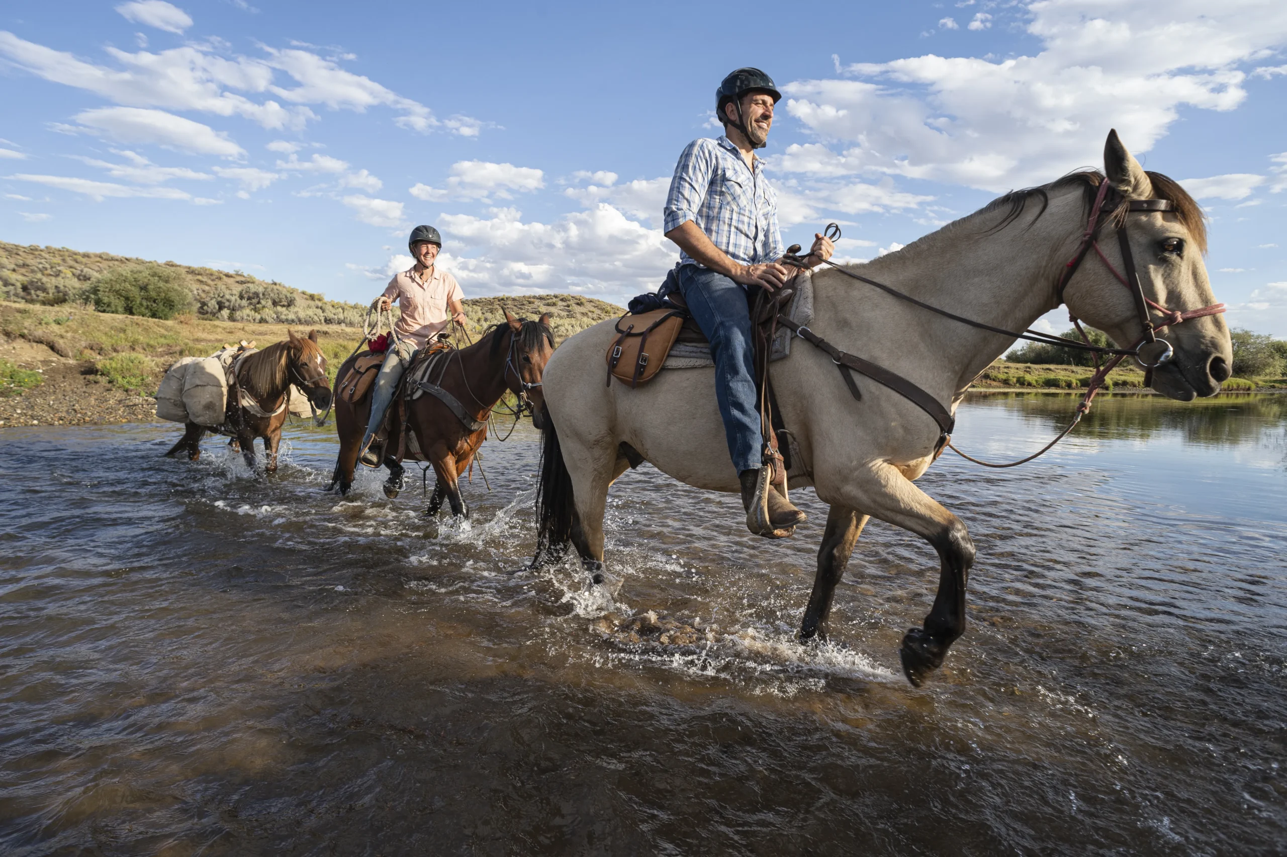 Two people on horseback cross a small river. Both people are smiling and wearing helmets