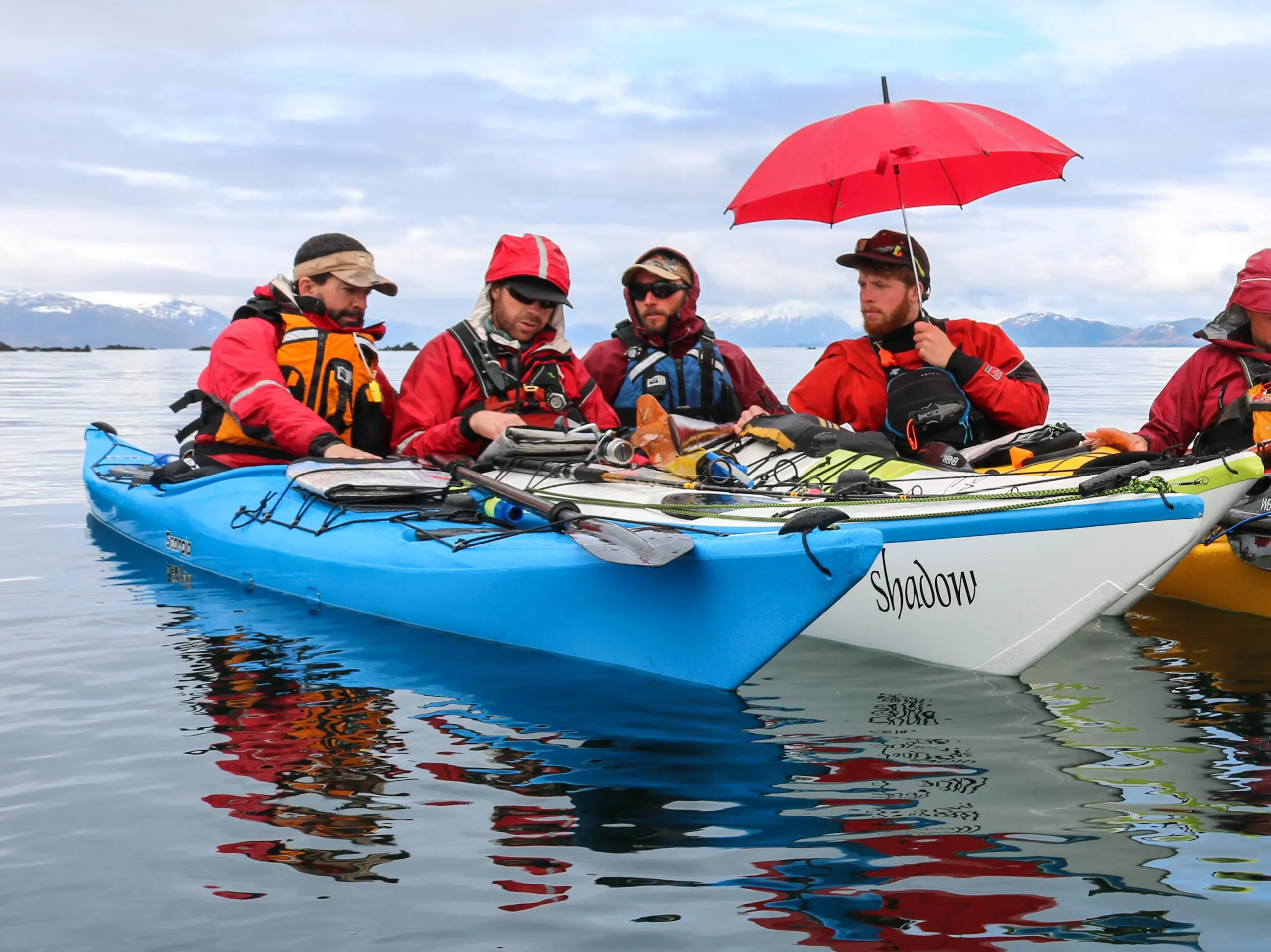 Four men in sea kayaks group together around a chart. The man on the right is holding a red umbrella to provide shade; the sky is blue.