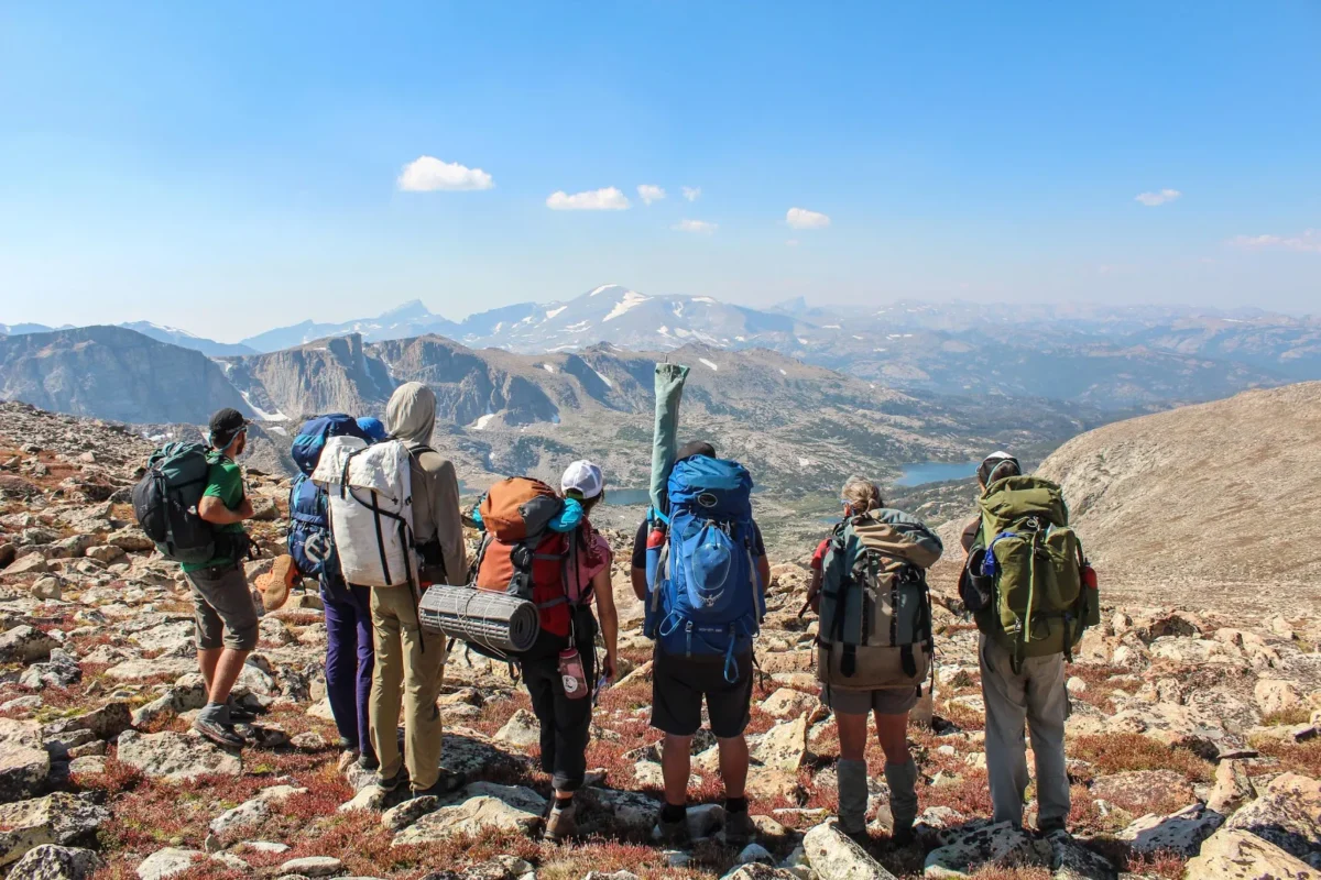 A group of people wearing backpacks of varying sizes stand on top of a mountain range, looking toward more mountains.