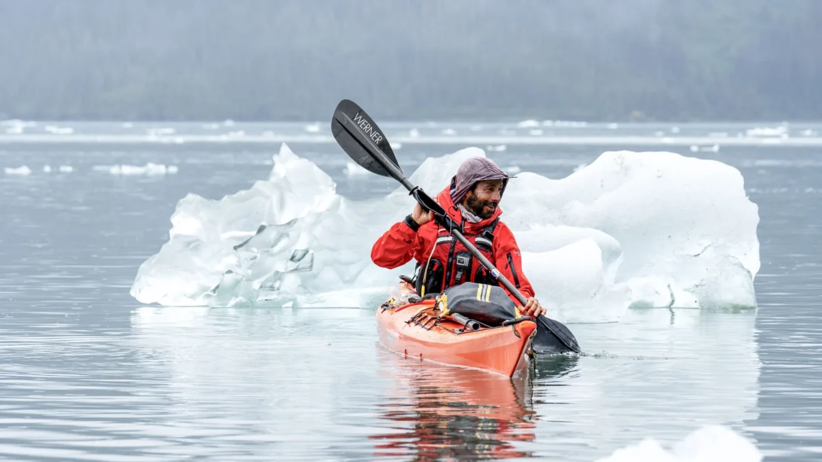 A sea kayaker paddles in a calm sea. Behind them is an iceberg.