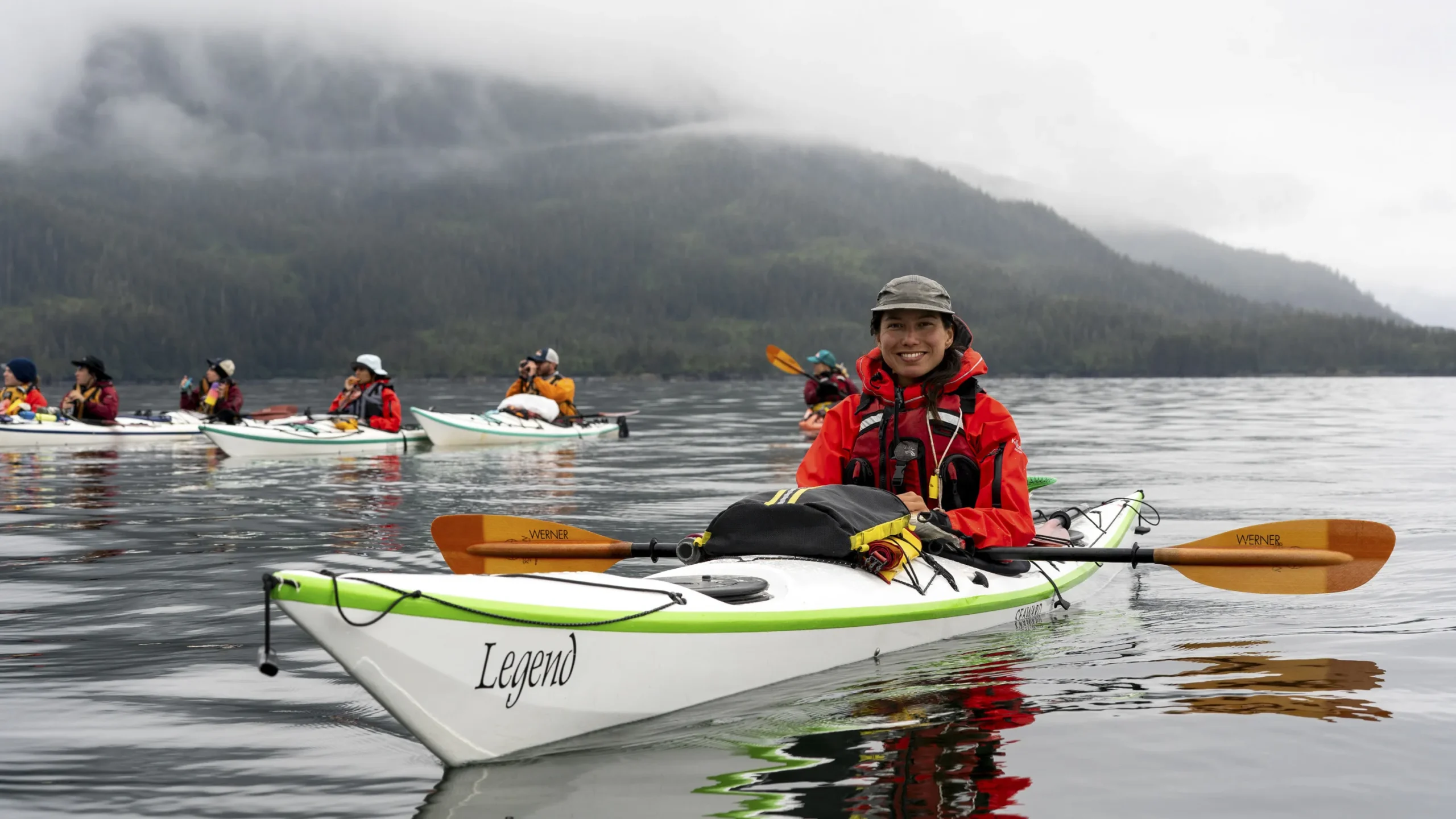 A group of sea kayakers on a flat ocean. There is a mountain shrouded in mist in the background.