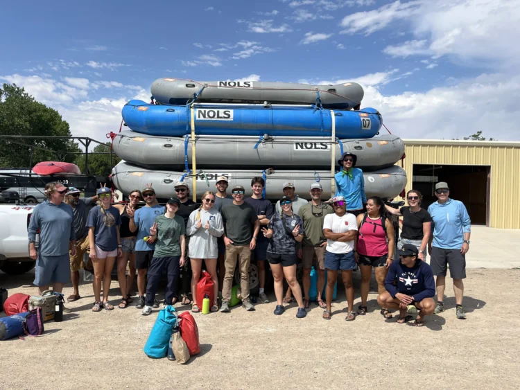 A large group of people pose in front of a trailer piled with river rafts. It's sunny and looks warm based on their clothing.