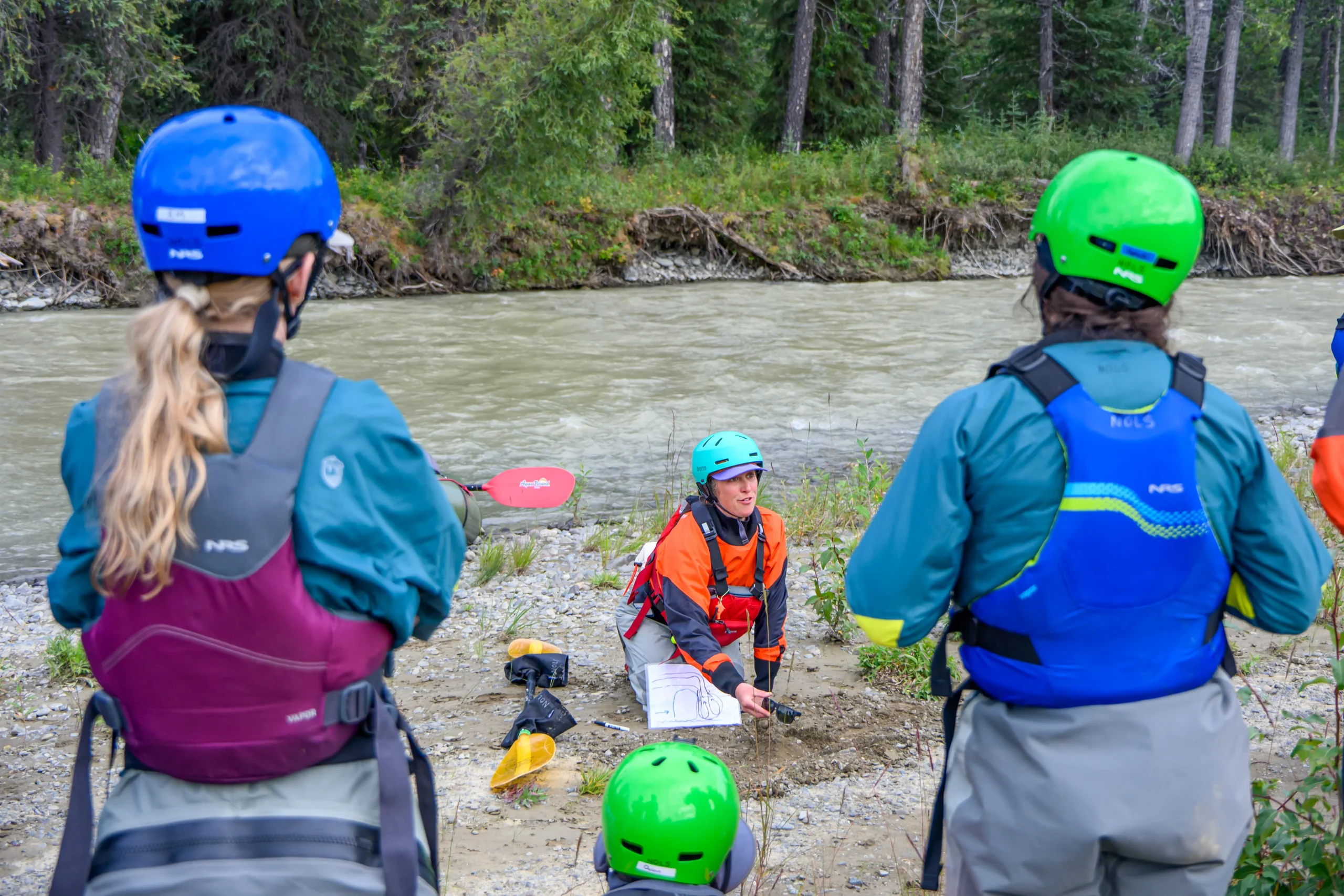 A woman in an orange drysuit and a yellow helmet sits on the bank of a river. She is pointing to a diagram. There are three people with wetsuits and helmets watching her.
