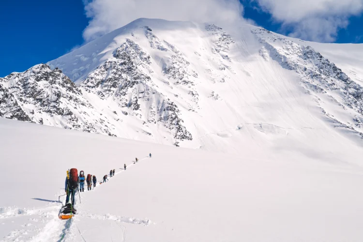 A line of mountaineers walks in single file through snow at the base of a large snowy peak