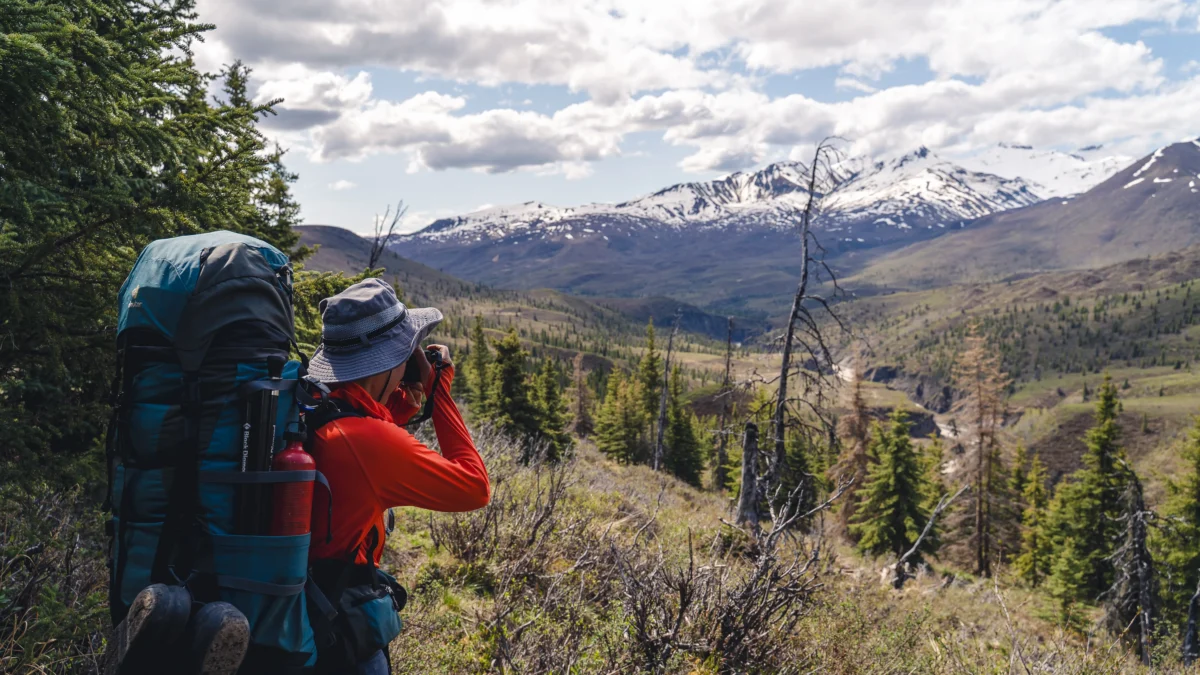 A backpacker uses binoculars to look at mountains in the distance
