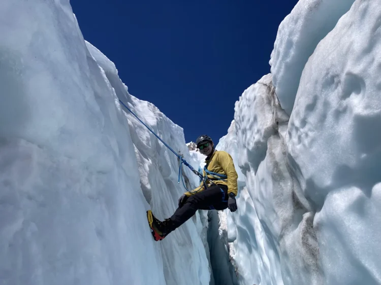 A person in a yellow jacket and black helmet rests their feet against a wall of ice. They are suspended by rope and harness in a crevasse.