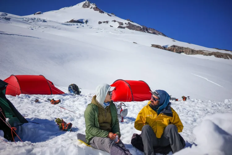 Two men sit in front of two red mountaineering tents. They are on the snow and there is snow and a mountain peak in the background. 