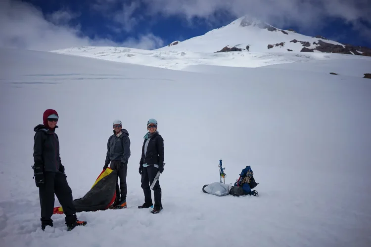 Three people stand on a snowfield assembling a tent. 