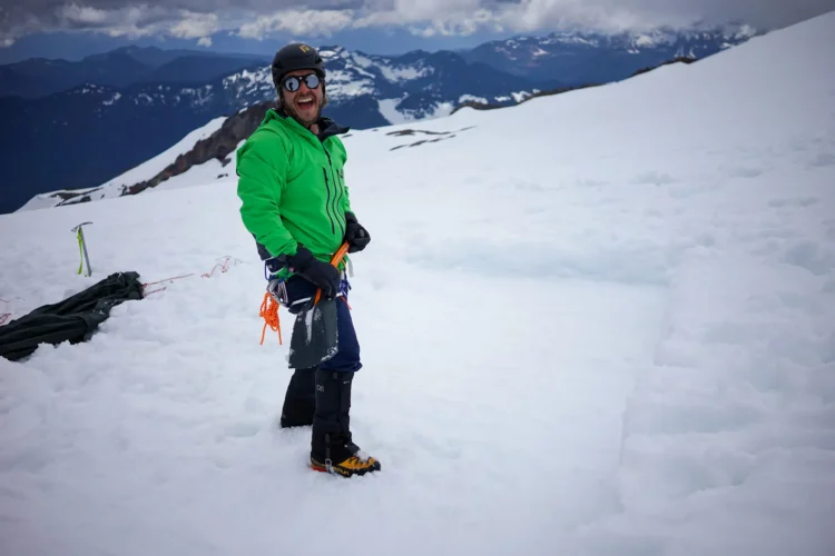 a man in a bright green jacket, helmet, and sunglasses smiles at the camera. He is standing on snow partway up a mountain. There are lots of snow-topped mountains in the background.