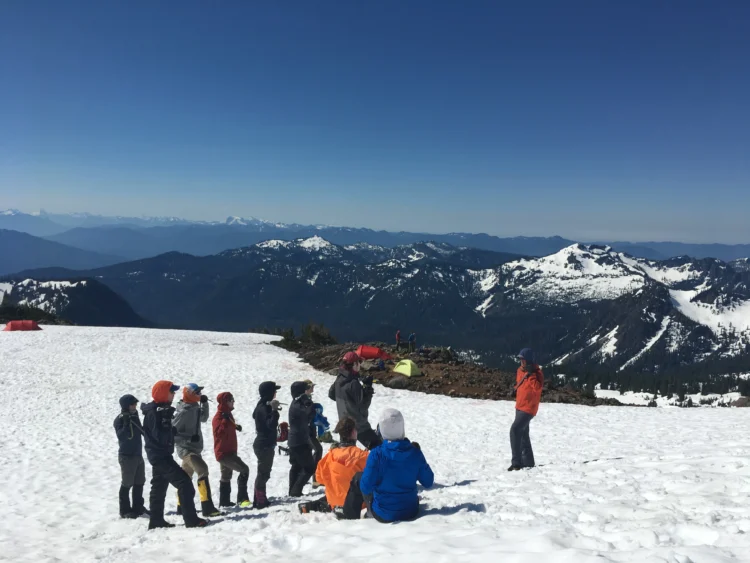 a group of people in technical outdoor gear stand in a group looking at someone who appears to be teaching. They are on the side of a mountains and there are lots of mountains in the background. 