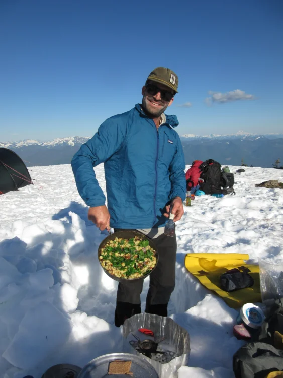 A man in a blue jacket poses on the snow with a pan of hot food. He's smiling.