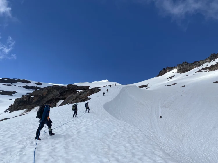Mountaineers in a rope team ascend a snow field between rock outcroppings. The sky is bright blue.