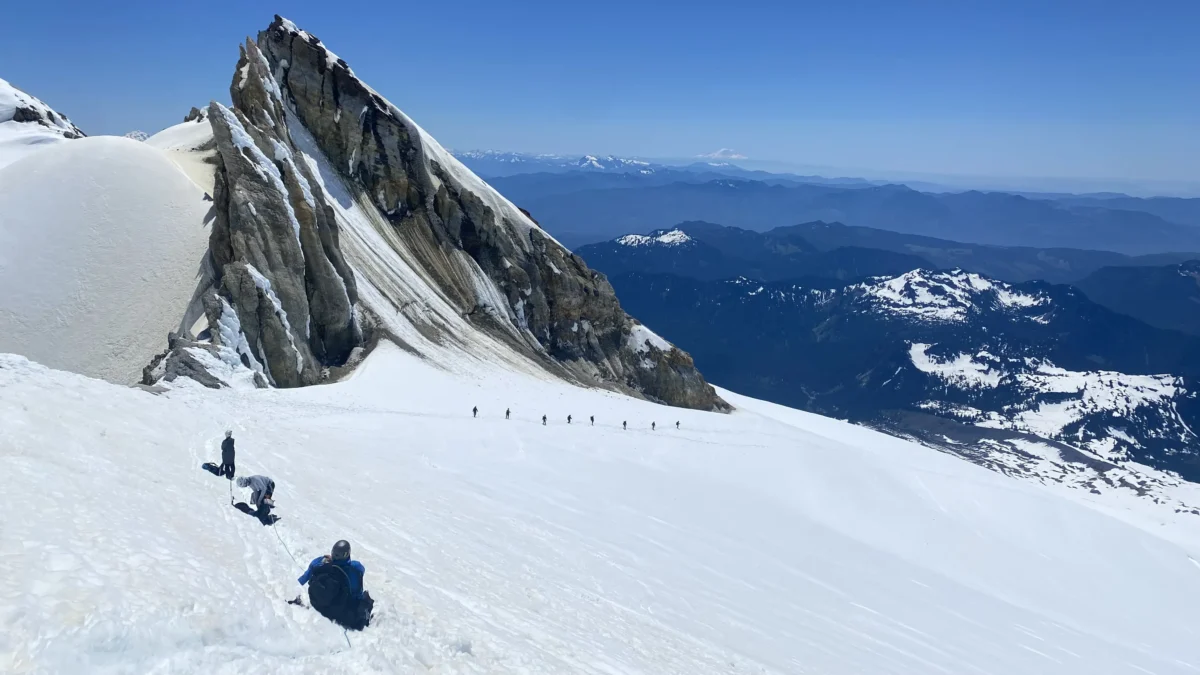 A line of climbers on snow snake beneath a giant fin of granite. In the background are more mountains