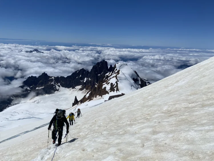 a group of mountaineers on a rope team ascend a snowy climb. There are clouds below them.