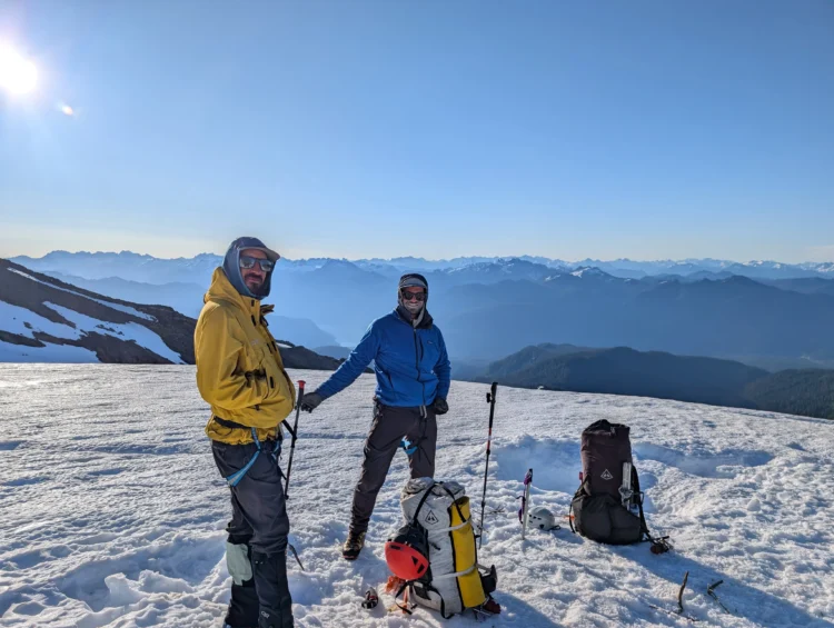 Two men pose partway up Mount Baker. They are standing on snow and there are mountains in the background.