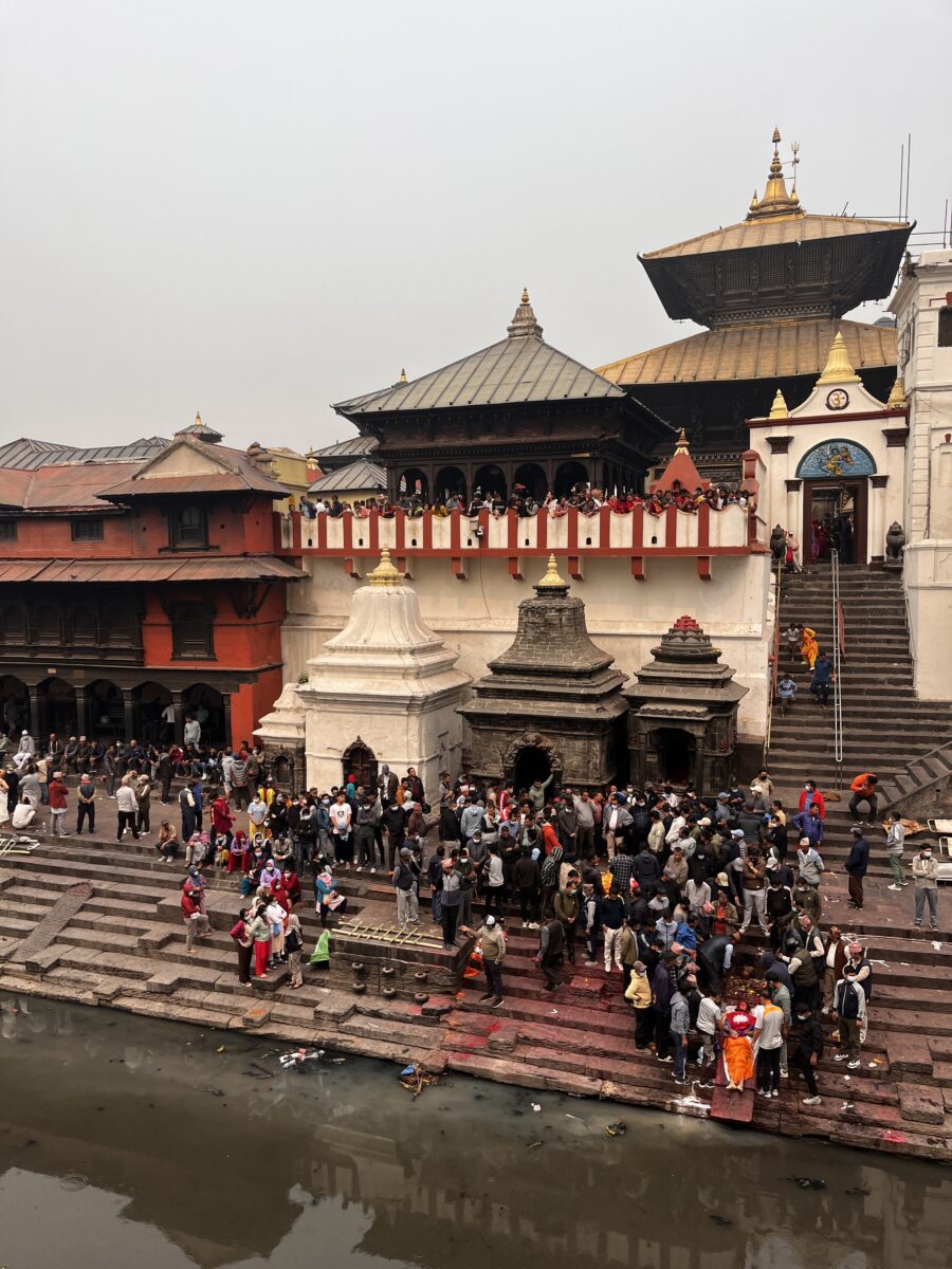 A street scene in Kathmandu. Crowds are in front of, and on the balcony of, an ornate building.