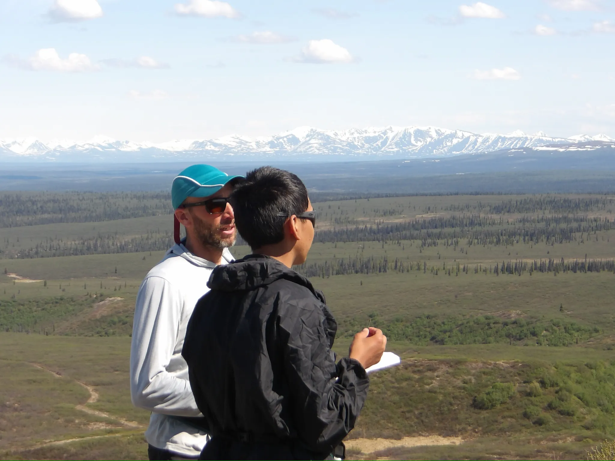 Two people look across a large green expanse at a range of snow-capped mountains in the distance.