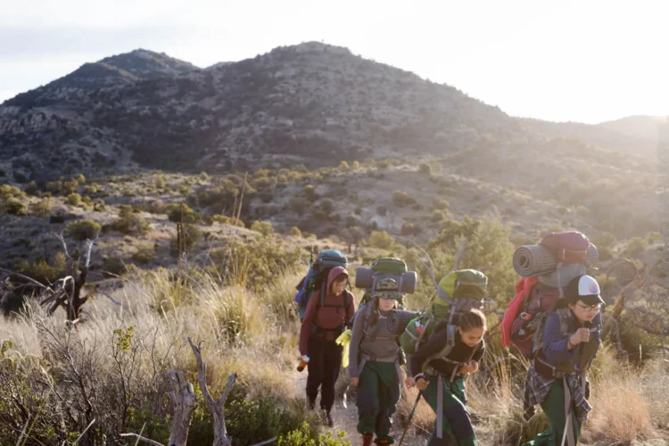 Group of teen backpackers hikes single-file through desert scrubland at golden hour, carrying full packs up a trail with arid mountains and scattered vegetation in the warm afternoon light.