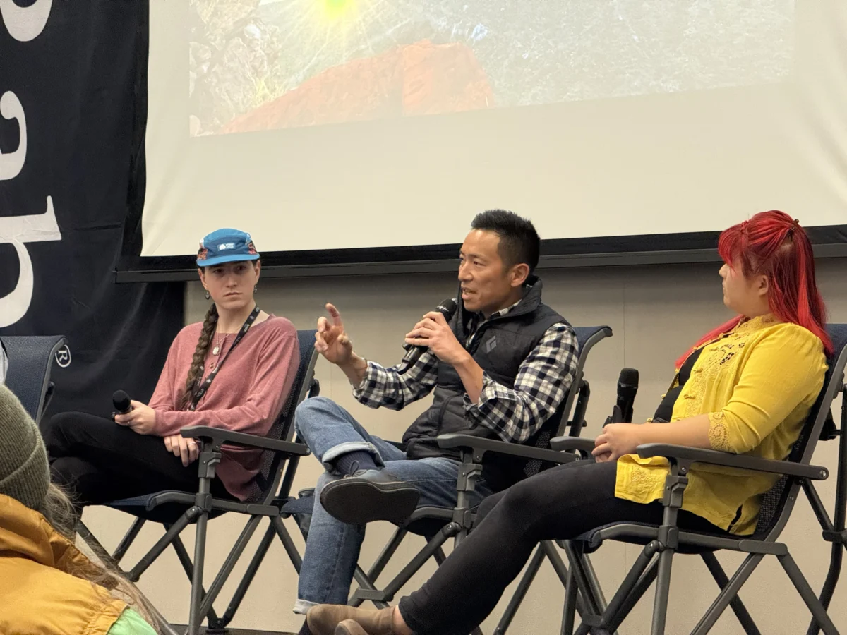 A man in a plaid shirt and black down vest sits with one leg folded across his knee. He is holding a mic and speaking. A person with red hair and a yellow sweater and a second person with a blue hat and long braid are watching him, seemingly engaged.