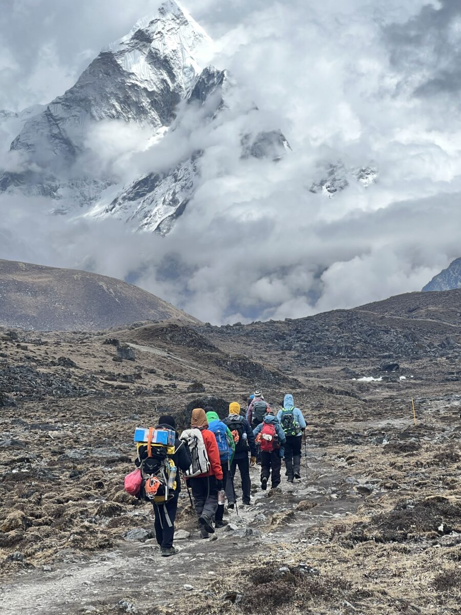 Team, wearing backpacks, walks toward Everest—shrouded in clouds—through bare and rocky terrain.