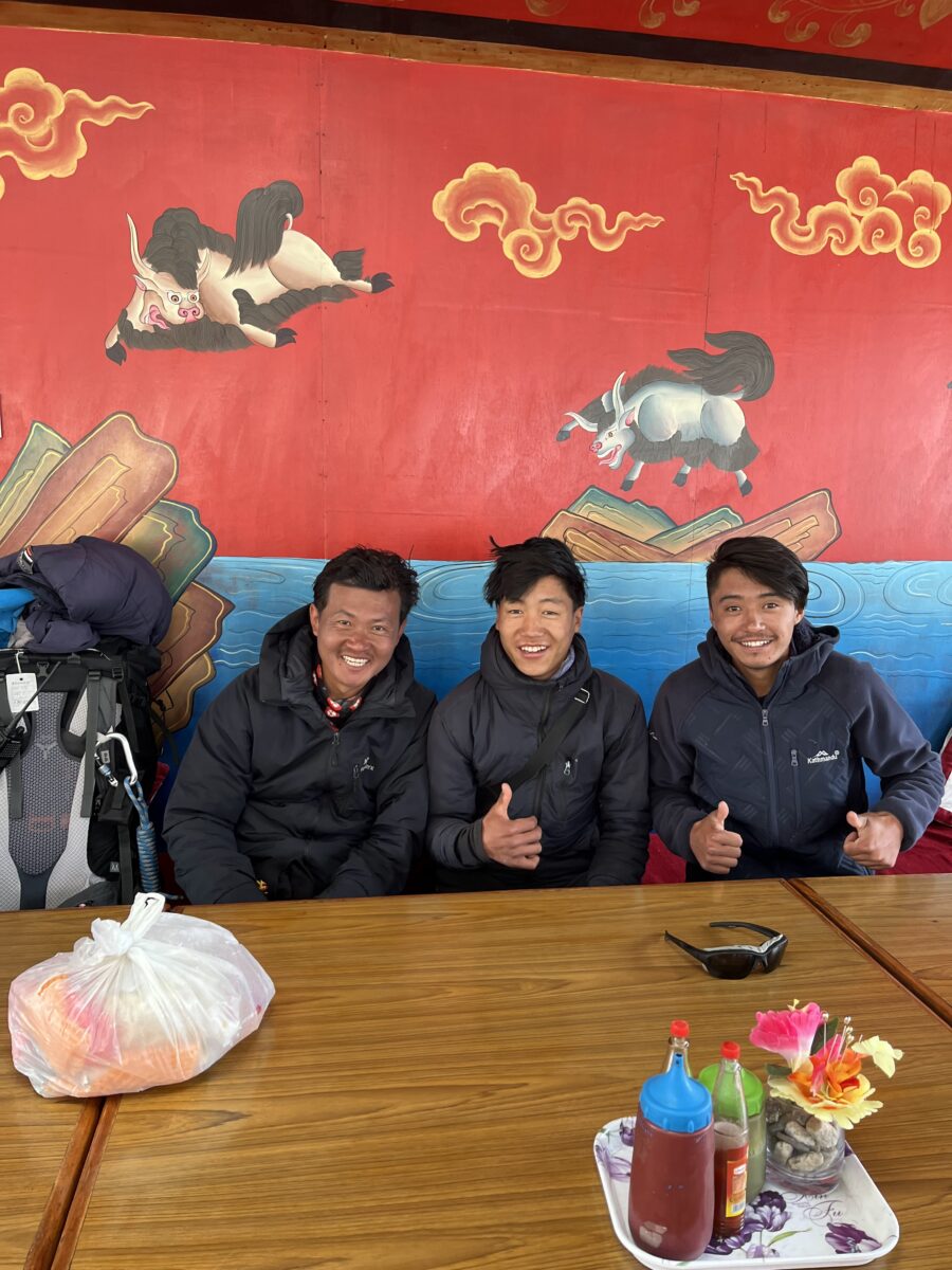 Three members of the team are photographed in a tea house in front of a brightly colored red and yellow painting.
