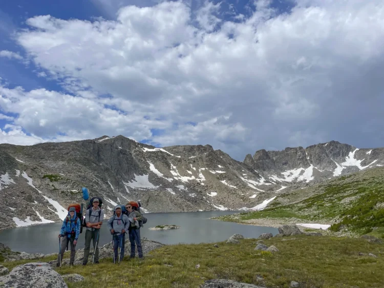 Four teen backpackers with large expedition packs stand at the edge of a lake in the Rocky Mountains, with snow-patched granite peaks and dramatic clouds reflected in the still water behind them.